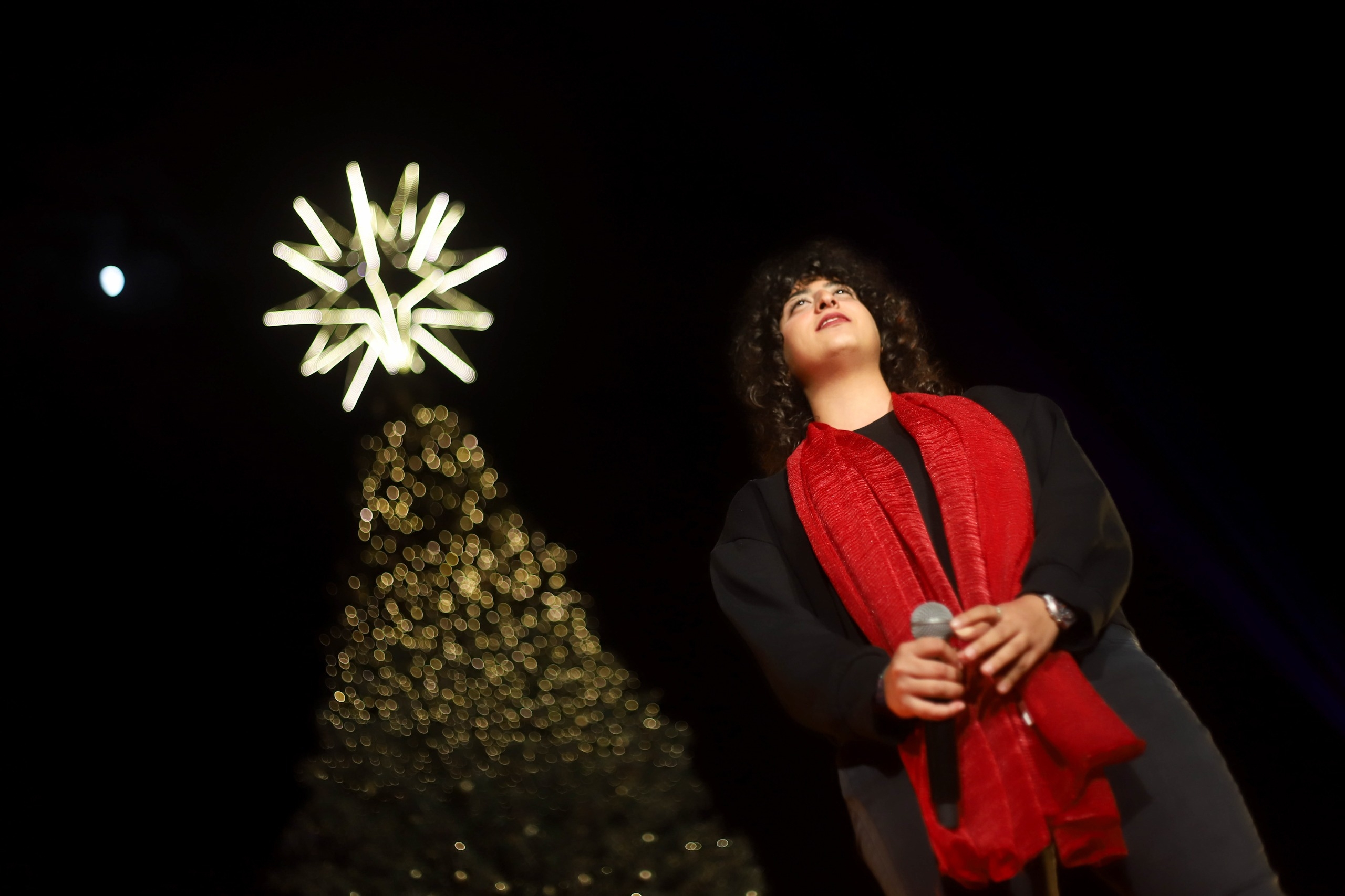 A girl singing in front of a Christmas tree adorned with lights and decorations, creating a festive holiday atmosphere.